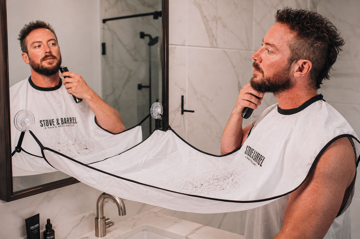 Man grooming beard with electric trimmer, wearing Stove & Barrel barber cape in modern bathroom