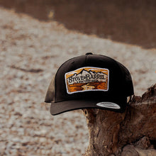 Black Stove & Barrel trucker hat with mountain logo, resting on rustic wood, embodying rugged modern masculinity.